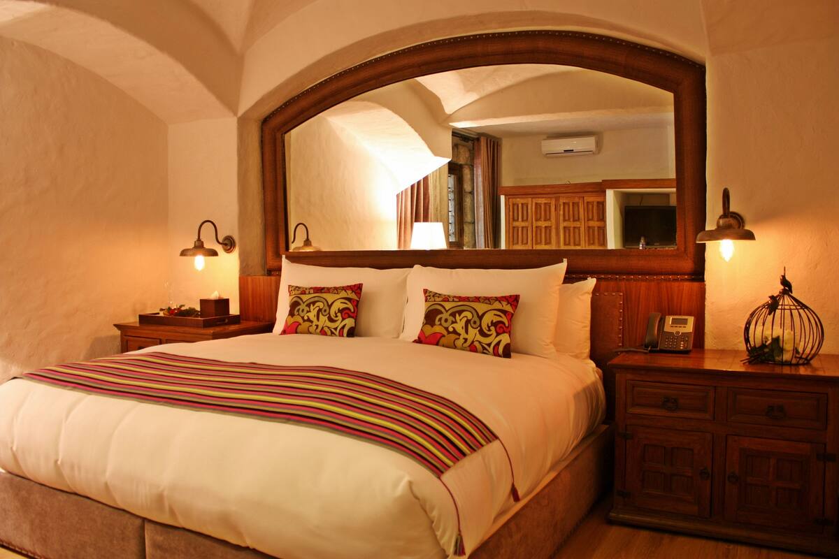 Een hotelkamer met een groot bed, een houten hoofdbord, een spiegel, bedlampen, een nachtkastje en een houten dressoir.