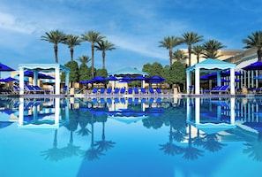 A pool area with blue umbrellas and lounge chairs, surrounded by palm trees and a clear sky.