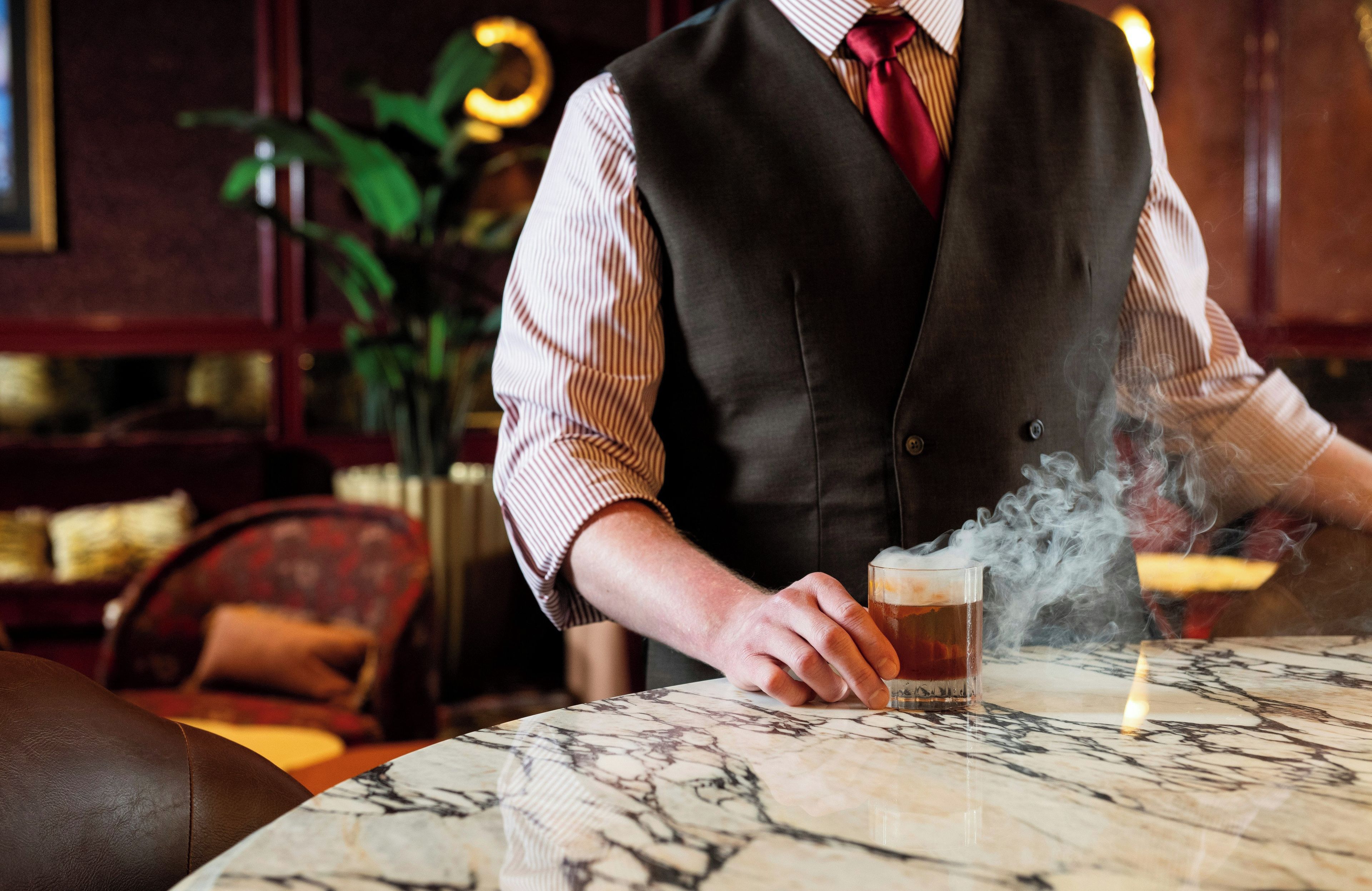 A person pouring a drink with steam rising from the glass, seated at a marble table in a room with a plant and cushioned chairs.
