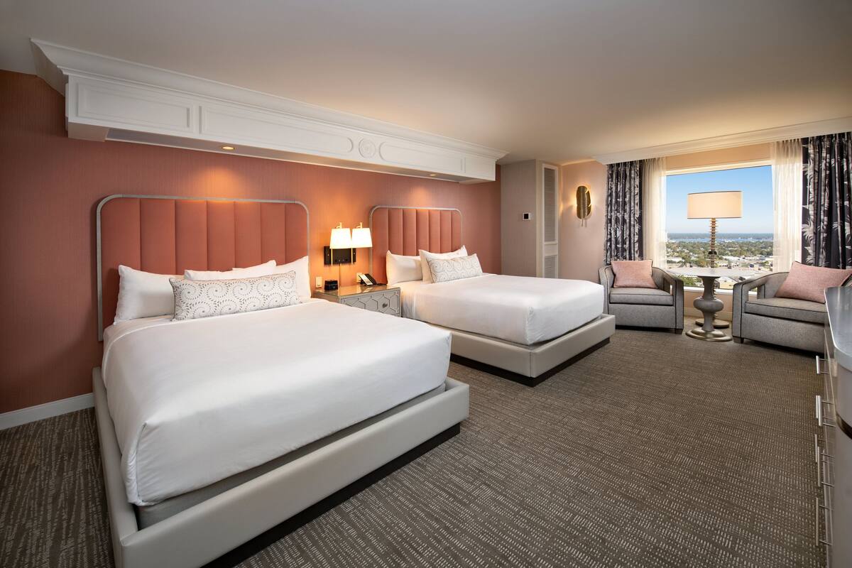 A hotel room with two beds, a sitting area with a table and chairs, and a view through a window.