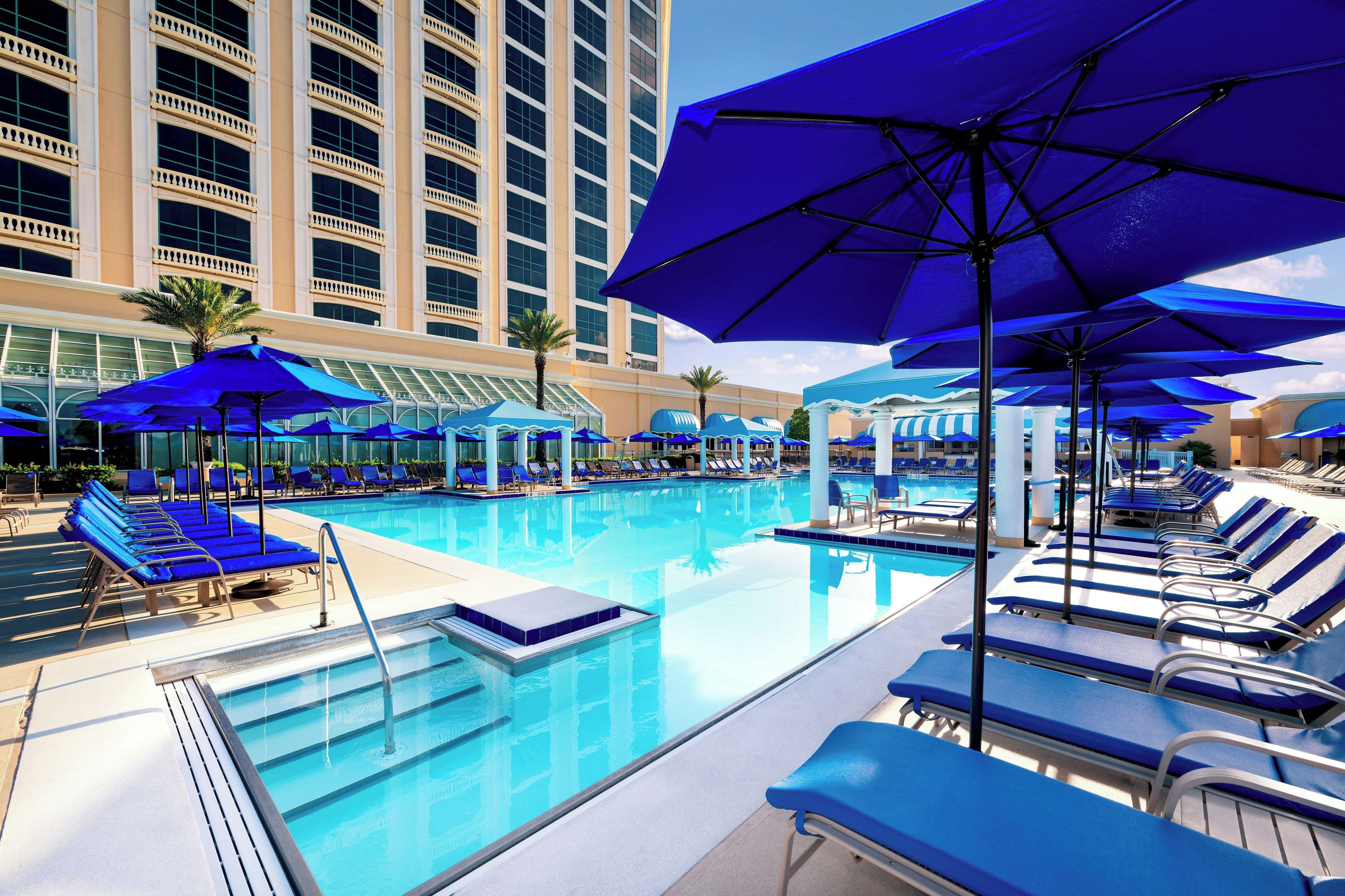 Uma área de piscina de hotel com várias espreguiçadeiras e guarda-sóis, uma piscina azul límpida e um edifício grande ao fundo.