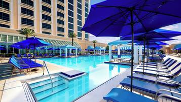 Uma área de piscina de hotel com várias espreguiçadeiras e guarda-sóis, uma piscina azul límpida e um edifício grande ao fundo.