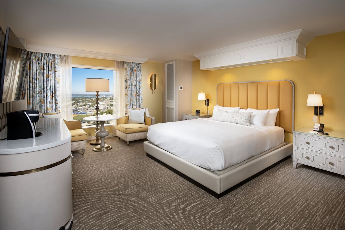 A hotel room with a large bed, a desk with a computer, a chair, a lamp, and a view of the city.