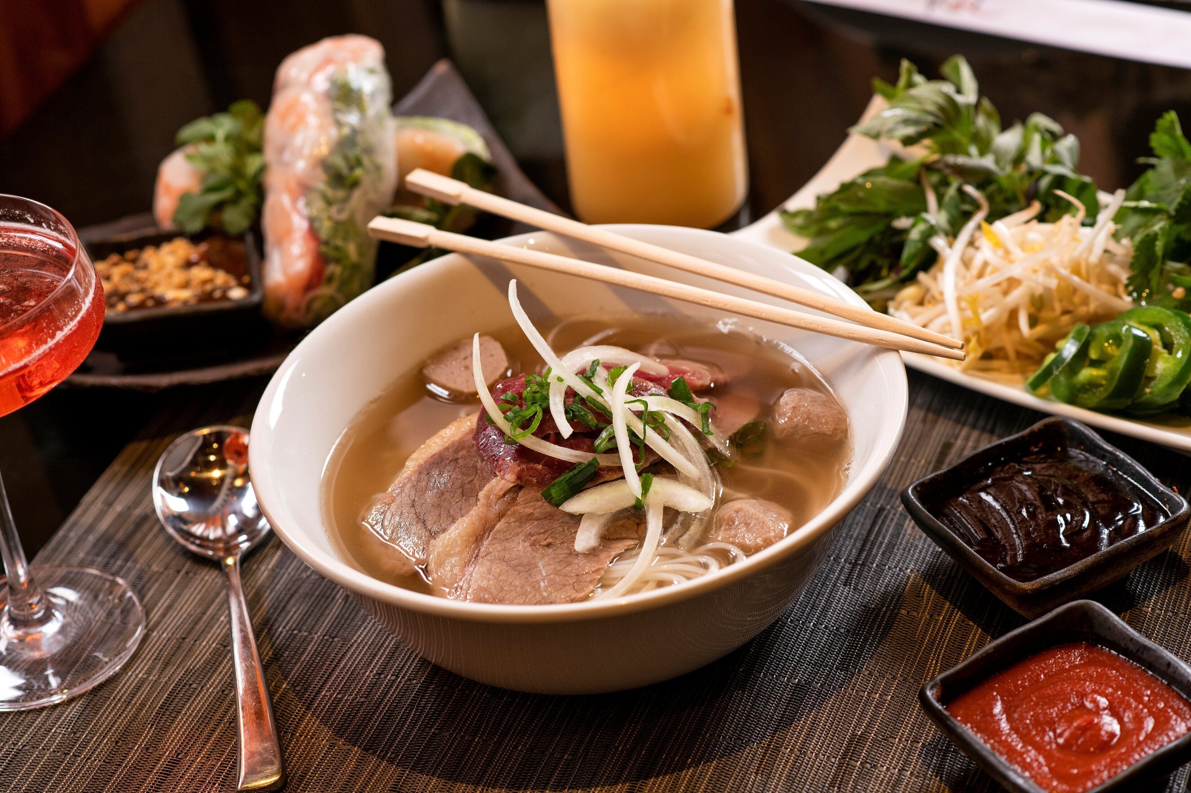 A bowl of soup with chopsticks, a spoon, and condiments on a table.