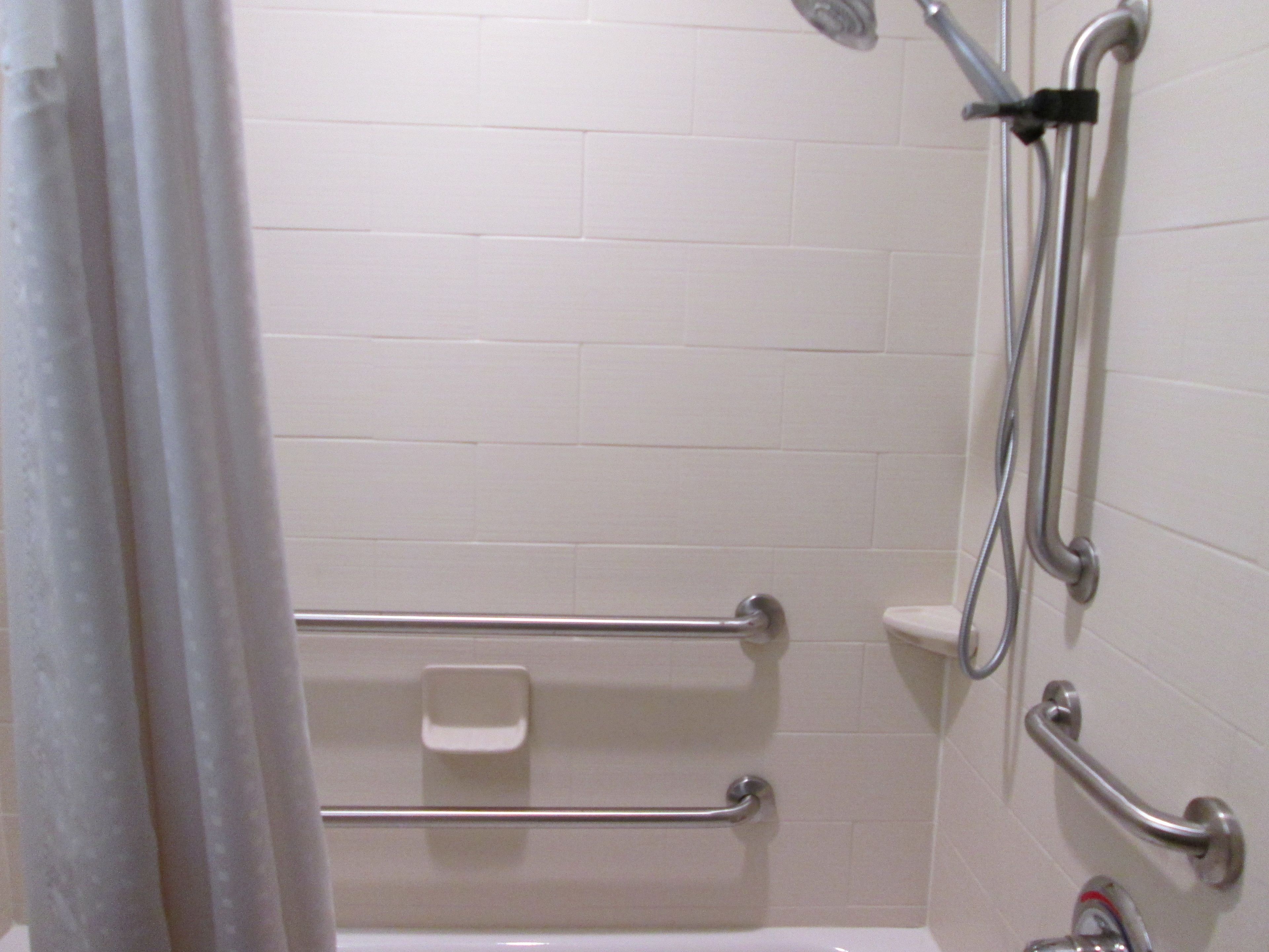 A bathroom with white tiled walls, a bathtub with grab bars, a handheld showerhead, and a handheld faucet.