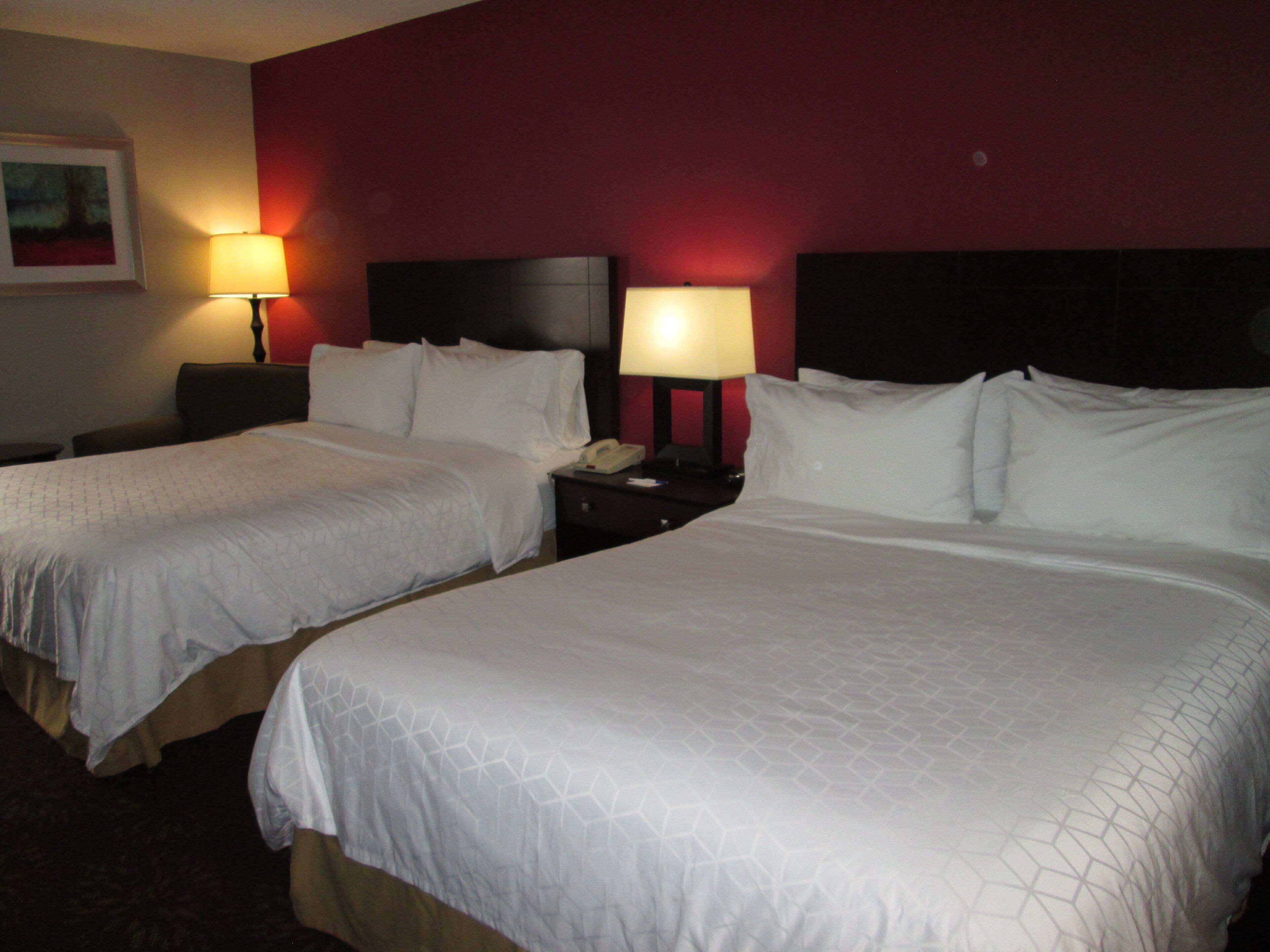 Una habitación de hotel con dos camas, cada una con ropa de cama blanca y cabeceros, una mesita de noche con lámpara y un cuadro en la pared.