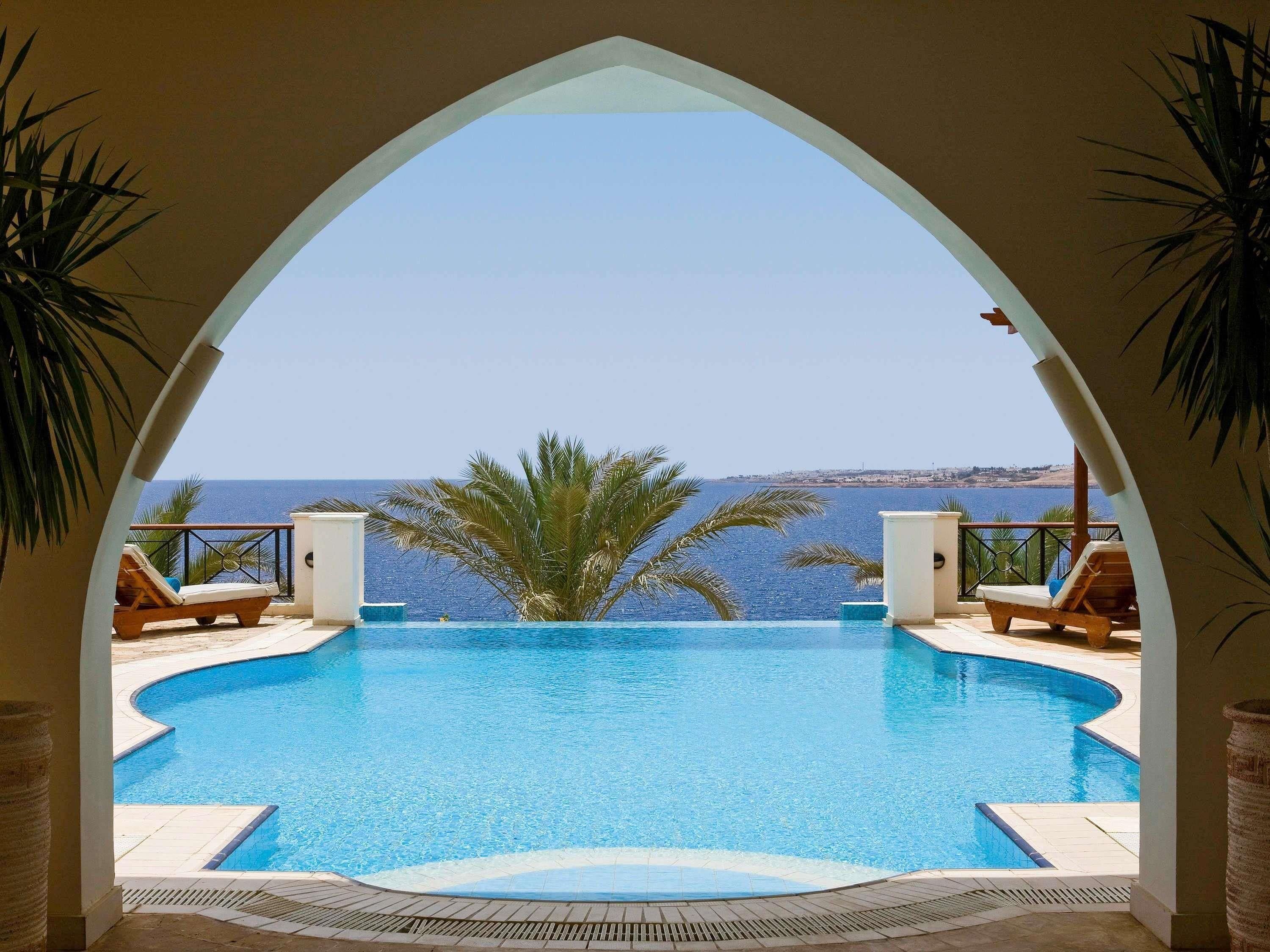 An infinity pool with a clear view of the sea, surrounded by lounge chairs and palm trees.