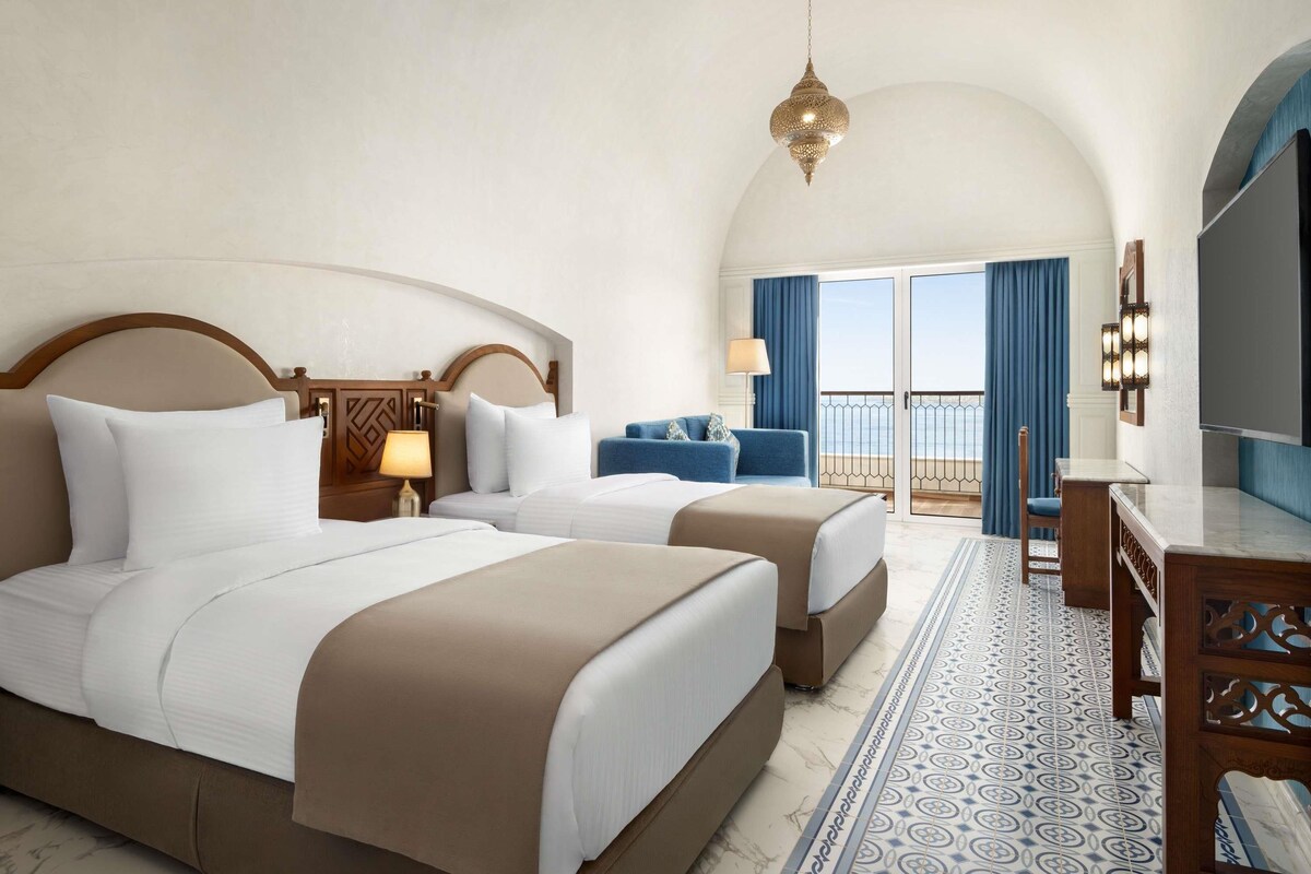 A hotel room with two beds, a desk, a TV, and a balcony with a view.