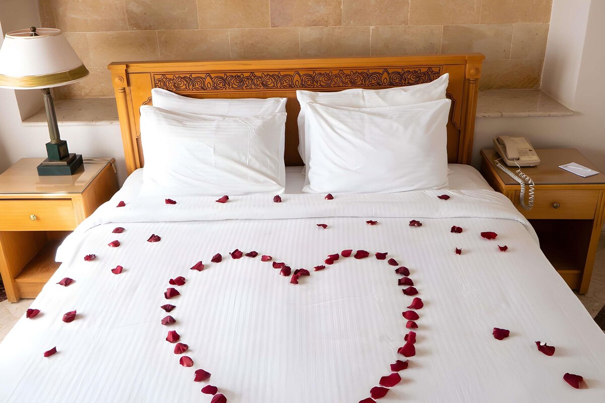 A hotel room with a bed, two pillows, a bedside lamp, a telephone, and a framed picture on the wall.