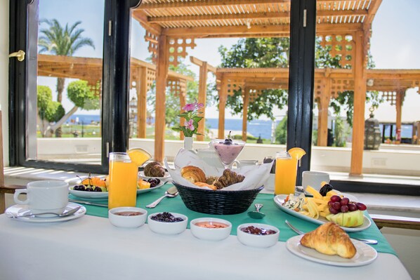 Una mesa de desayuno con diferentes platos, entre ellos frutas, pastelería y bebidas, con vista a una pérgola de madera y palmeras.