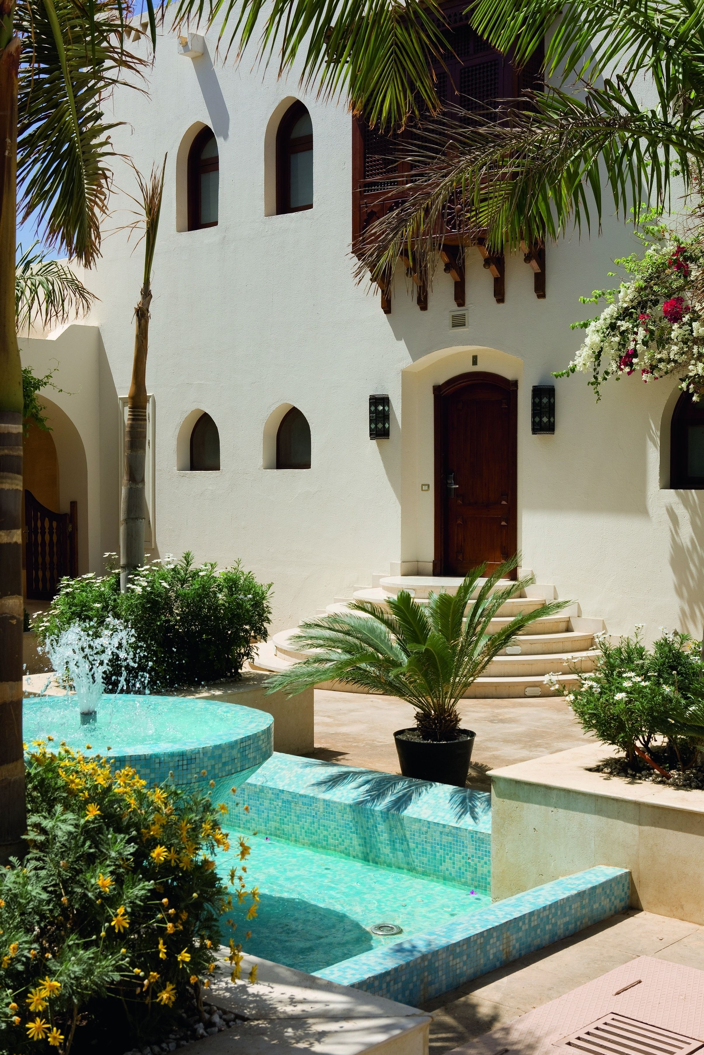 Un patio con piscina, palmeras y un edificio con ventanas de arco y puerta de madera.