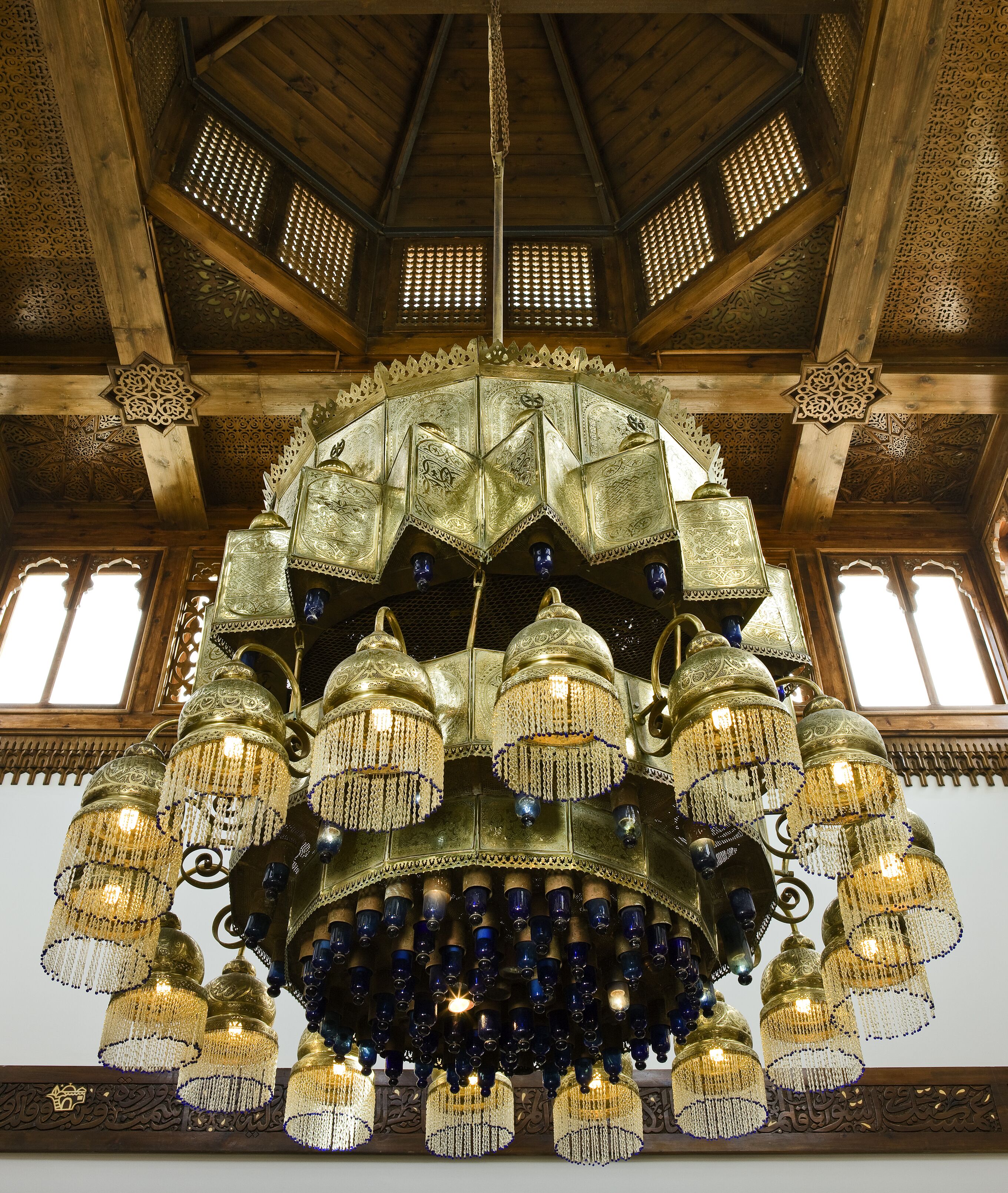 Un grande lampadario decorato con più livelli e luci pendenti, sospeso da un soffitto in legno con elaborate decorazioni.