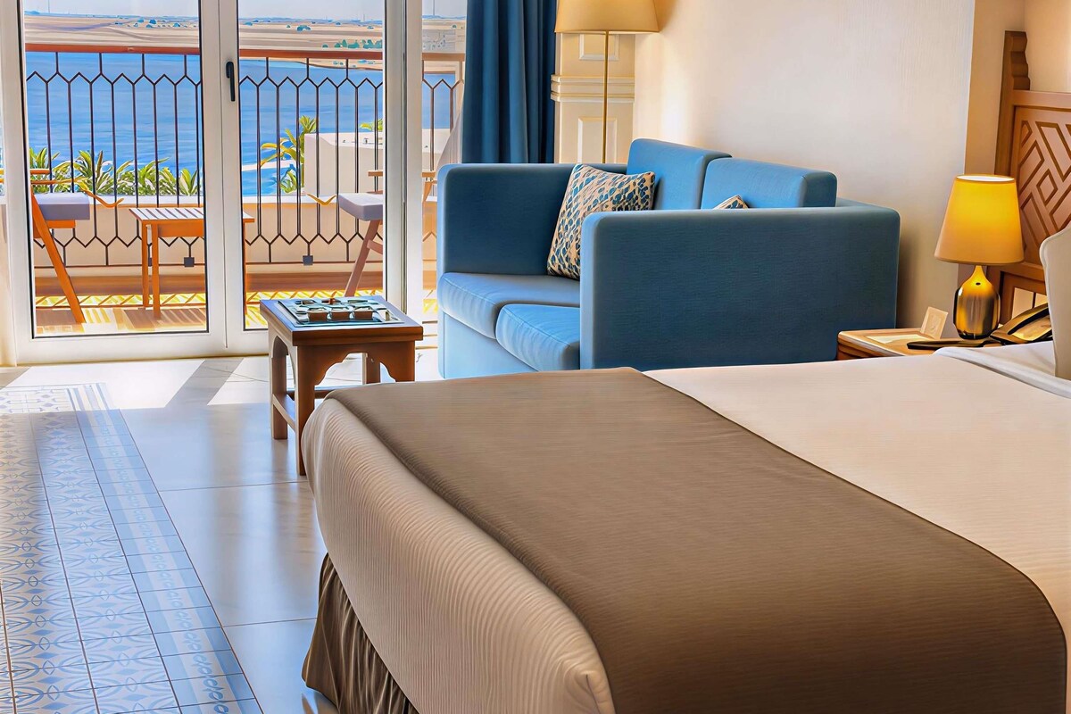 Una habitación de hotel con cama, sofá, una mesita y vista al mar a través de un ventanal.