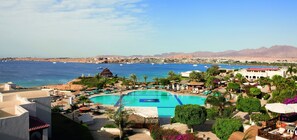 A resort with a large pool, palm trees, and a view of the sea and mountains.
