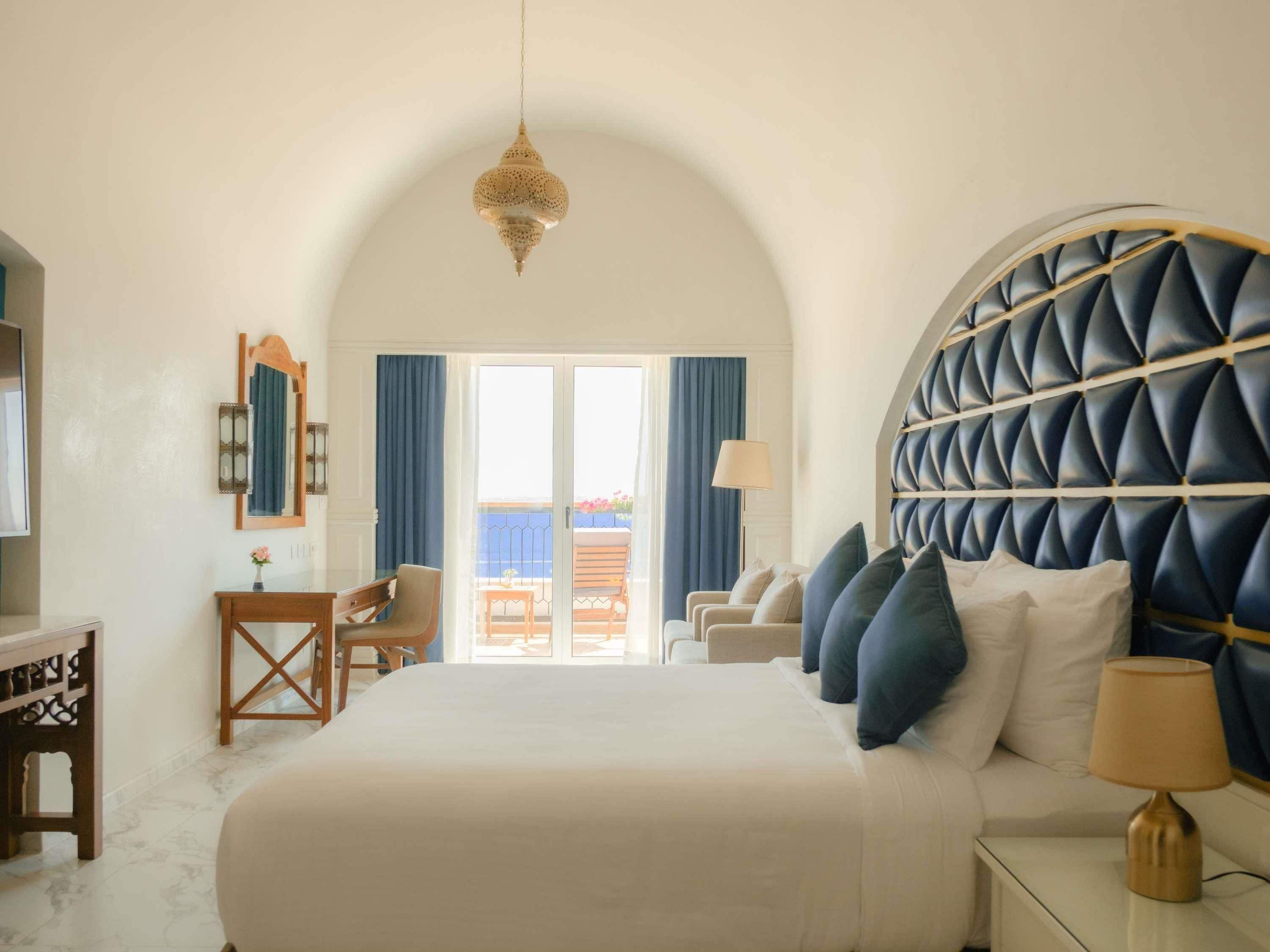 A bedroom with a large bed, a desk, a chair, and a view of the outside balcony.