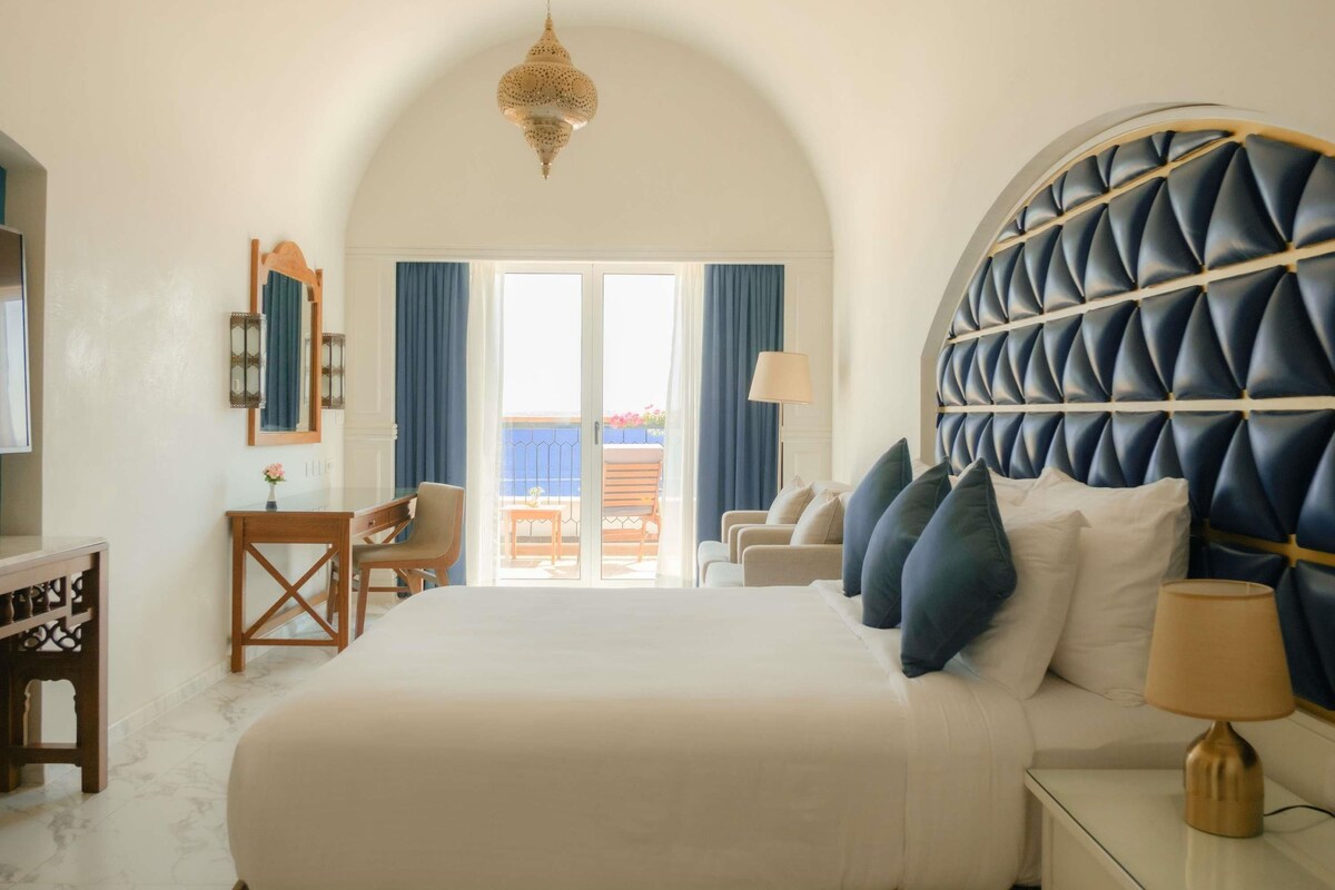 A bedroom with a large bed, a desk, a chair, and a view of the outside balcony.