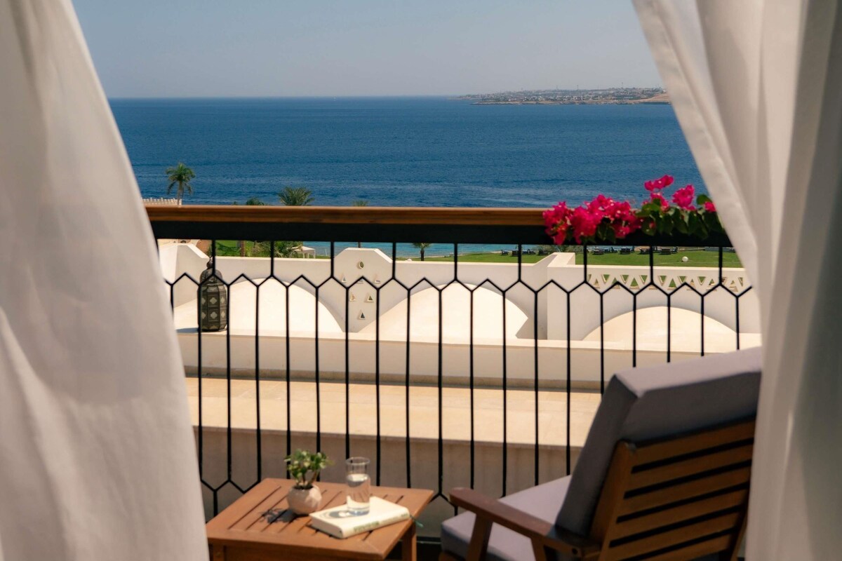Un balcón con vistas al mar, una mesa con un libro y un vaso, y una silla.