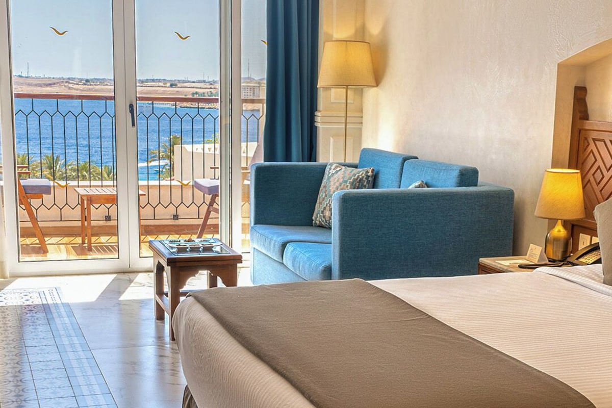 Habitación de hotel con una cama grande, un sofá, una mesita y vistas al mar a través de una puerta de vidrio.