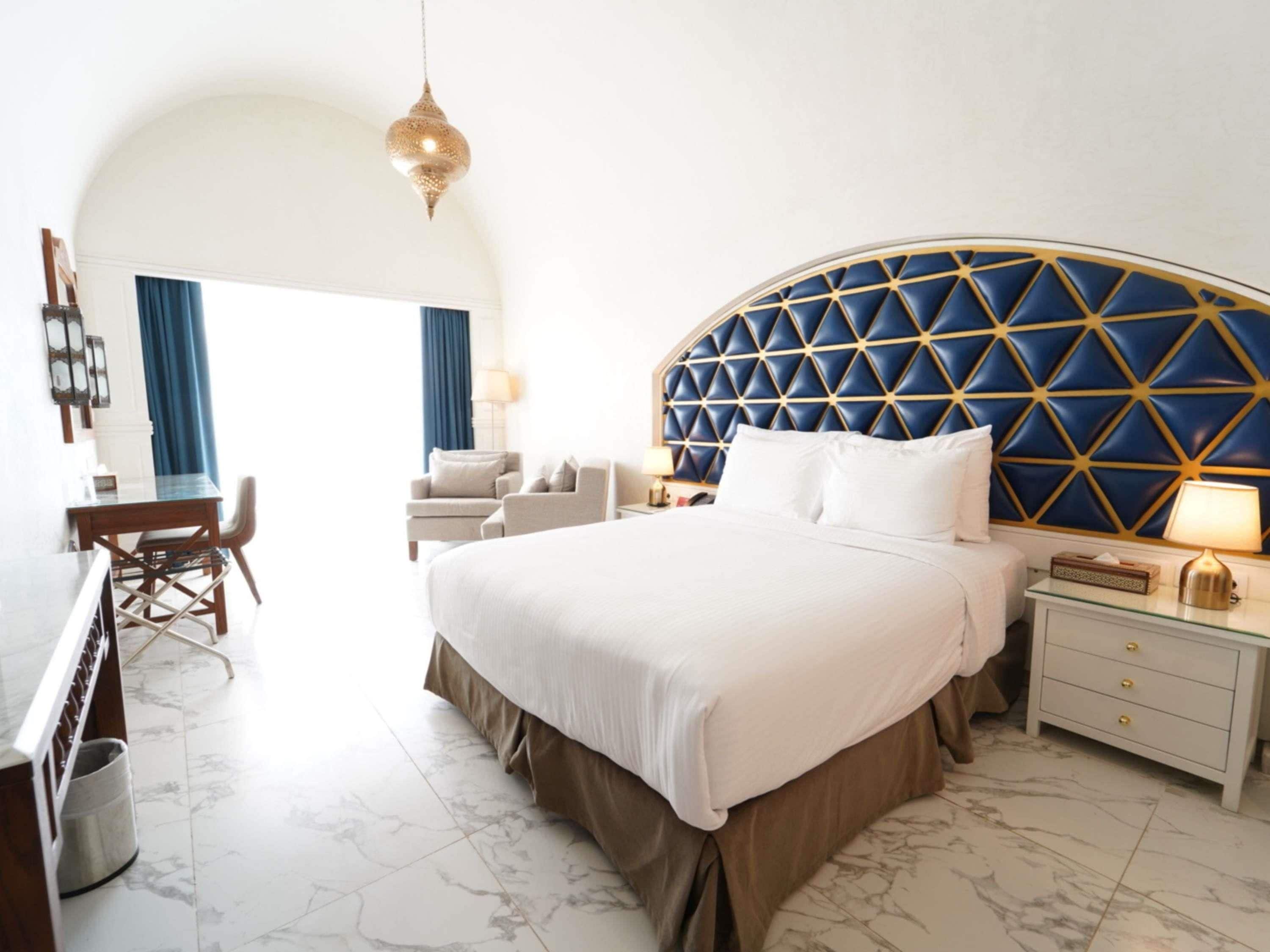 A hotel room with a large bed, a desk, and a chair. There is a blue and gold headboard, a hanging light fixture, and a nightstand with a lamp.