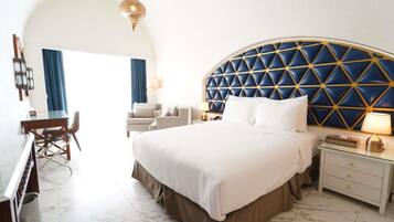 A hotel room with a large bed, a desk, and a chair. There is a blue and gold headboard, a hanging light fixture, and a nightstand with a lamp.