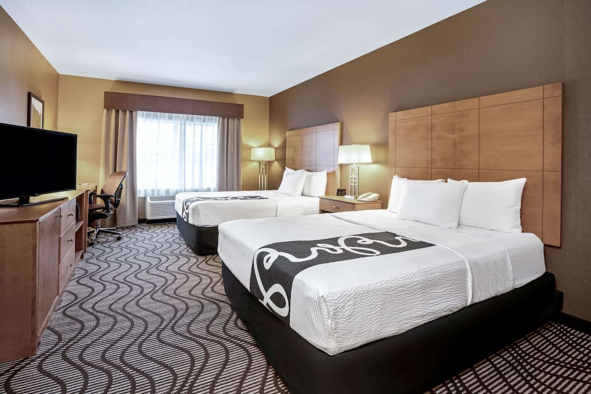 A hotel room with two beds, a TV, a desk, and a large window with curtains.