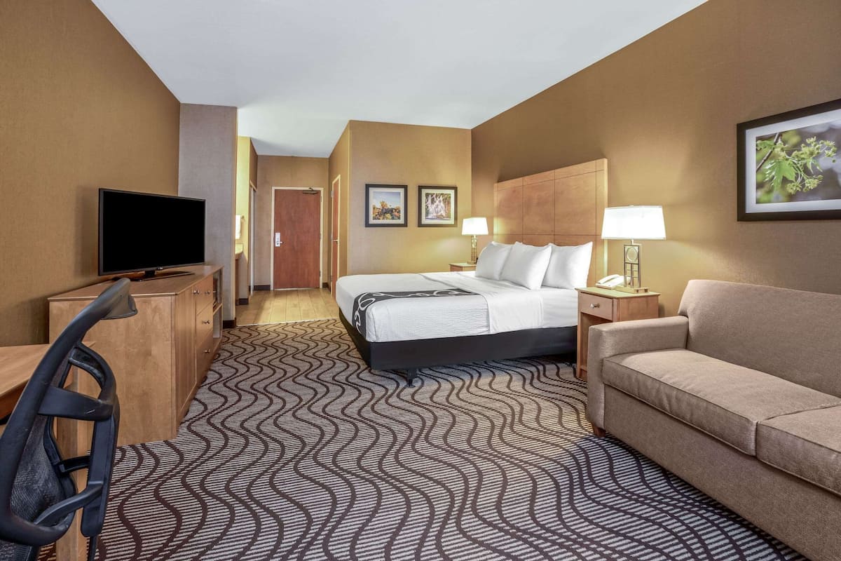 A hotel room with a bed, a desk with a chair, a TV, a sofa, and two bedside lamps.