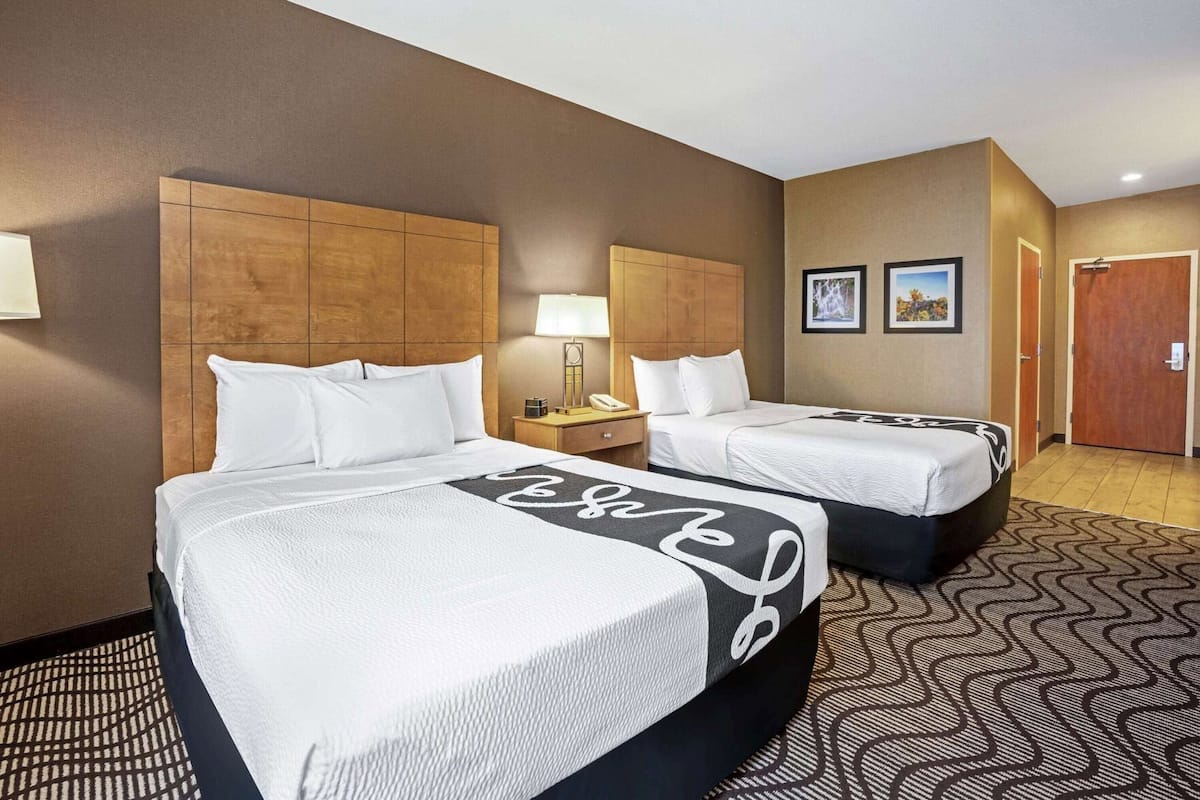 A hotel room with two beds, a wooden headboard, a nightstand, a lamp, and a door.