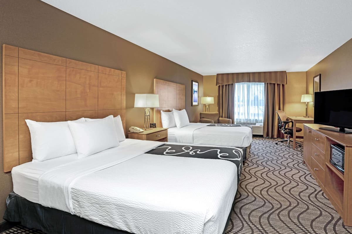 A hotel room with two beds, a desk, a television, and a large window with curtains.