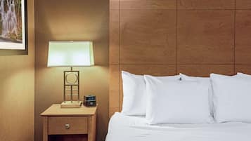 A neatly made bed with white linens and pillows, a bedside table with a lamp and a clock, and a framed picture on the wall.