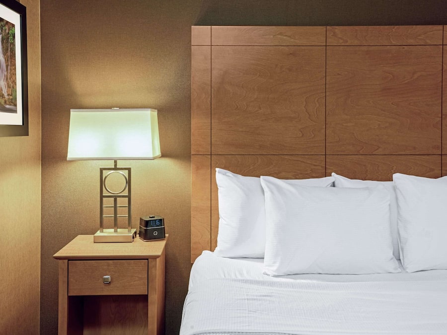 A neatly made bed with white linens and pillows, a bedside table with a lamp and a clock, and a framed picture on the wall.