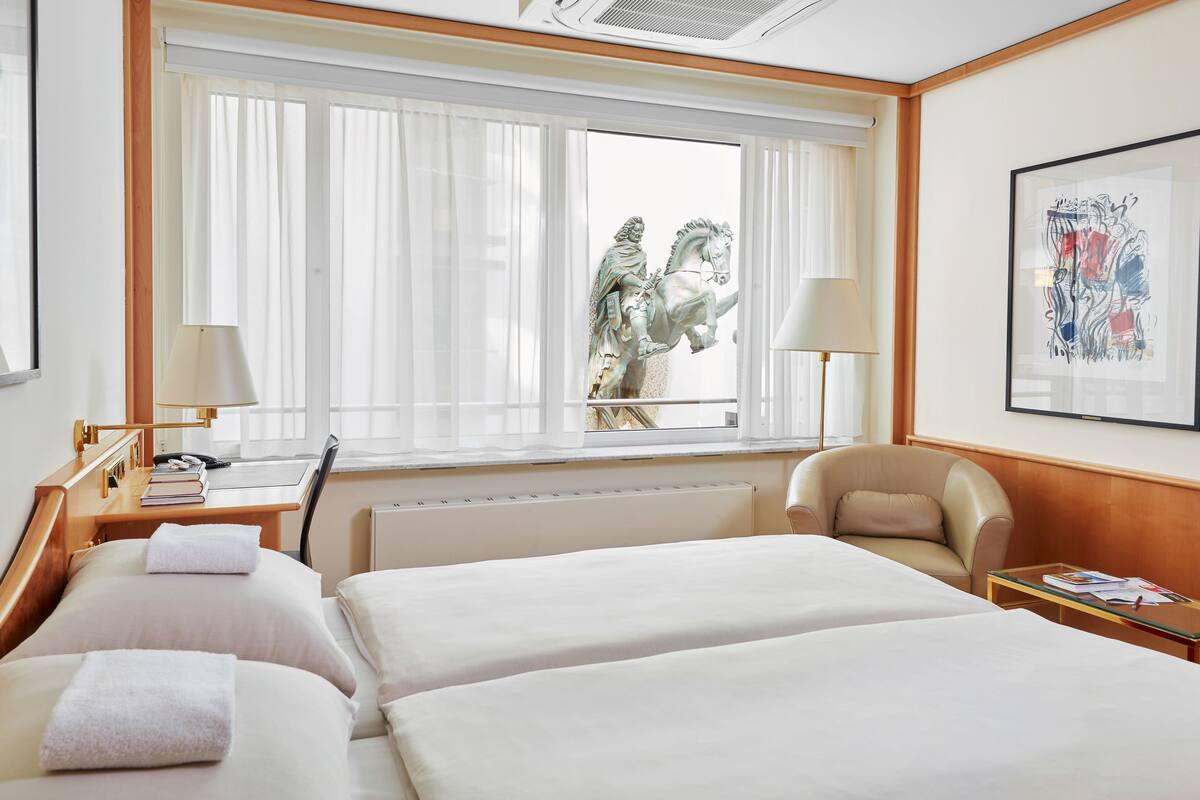 A hotel room with two beds, a desk with a lamp, a chair, a radiator, and a window with curtains.
