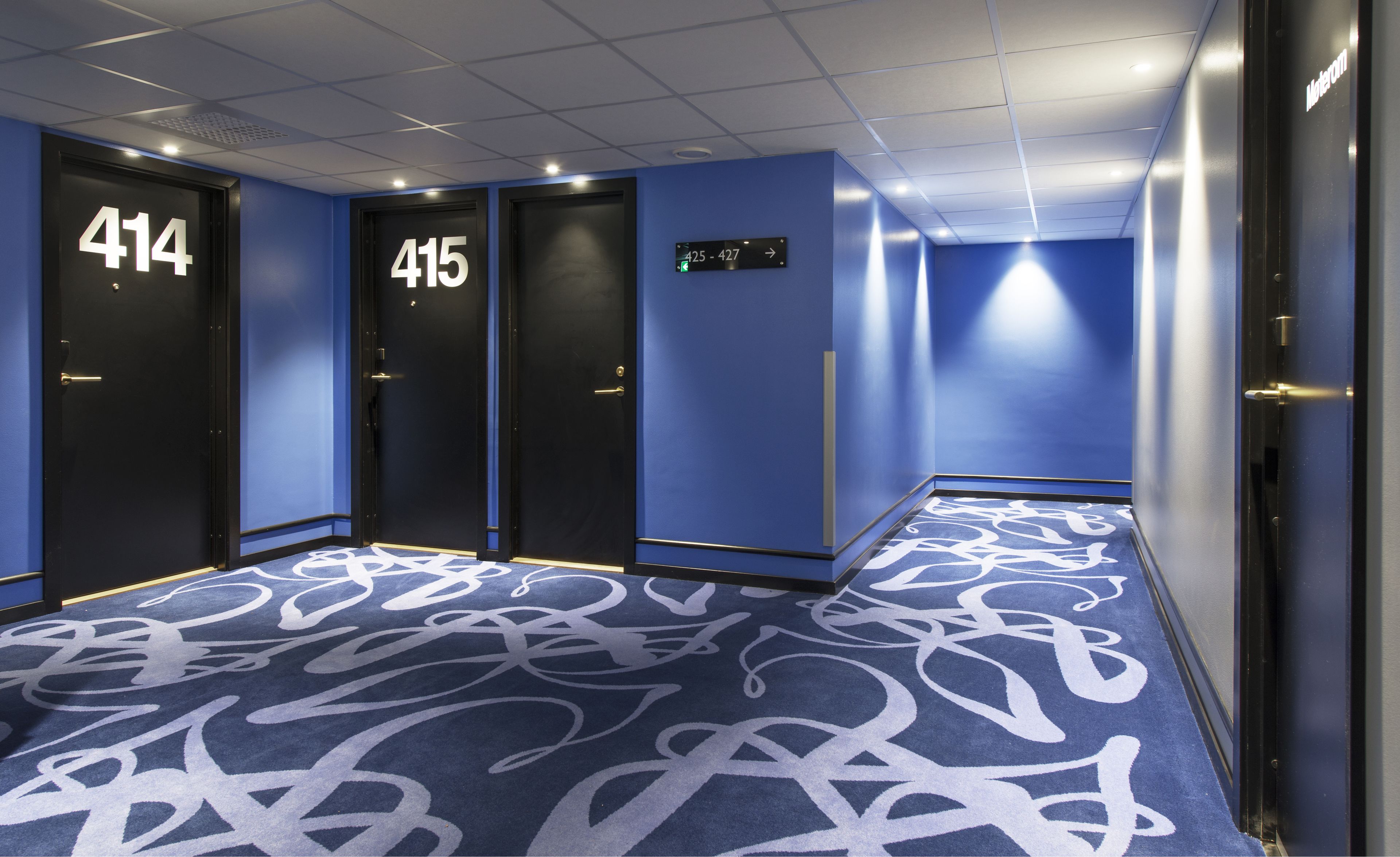 A hotel hallway with doors numbered 414 and 415, a blue carpet with white patterns, and a sign indicating room numbers 425-427.
