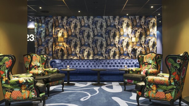A room with a blue sofa, two chairs with fruit patterns, a small table, and a wall with a peacock feather pattern. - Thon Hotel Terminus (Oslo)
