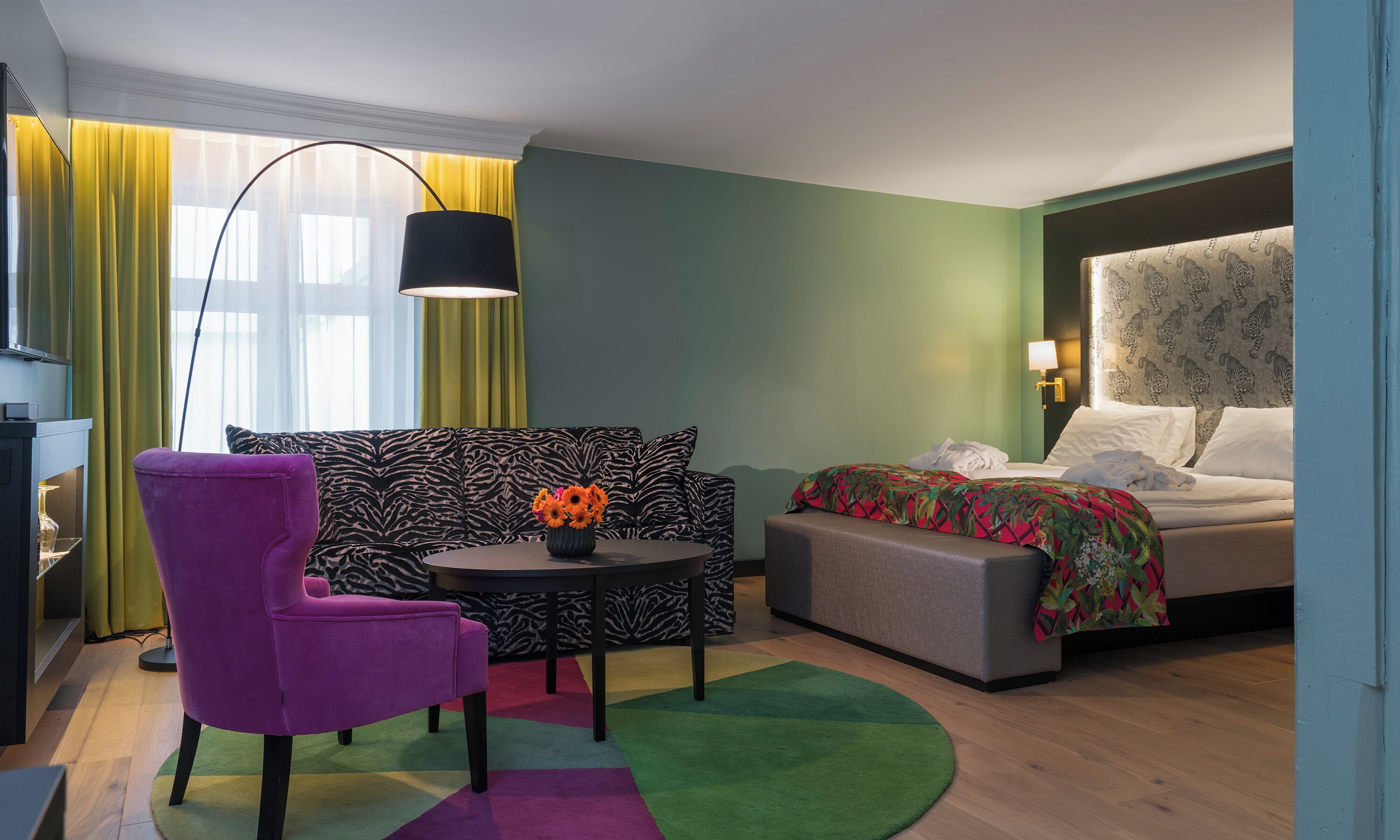 A hotel room with a zebra-patterned sofa, a purple armchair, a small round table with a vase of flowers, a bed with a colorful blanket, and a large floor lamp.