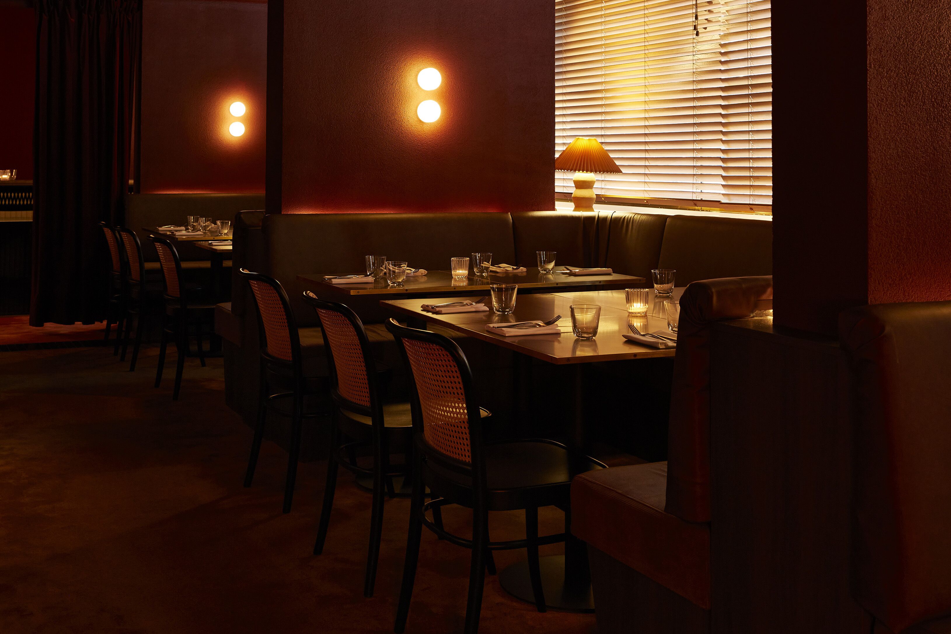 Un restaurant à la lumière tamisée, avec une longue table, plusieurs chaises et une fenêtre équipée de stores.