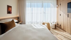 Quarto de hotel com uma cama grande, mesa de cabeceira, abajur, guarda-roupa de madeira e janela com cortinas translúcidas.