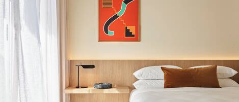 A hotel room with a bed, a nightstand, a lamp, and a modern abstract artwork on the wall.