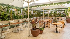 Um café ao ar livre com mesas e cadeiras, rodeado por grandes vasos de plantas e um toldo verde.
