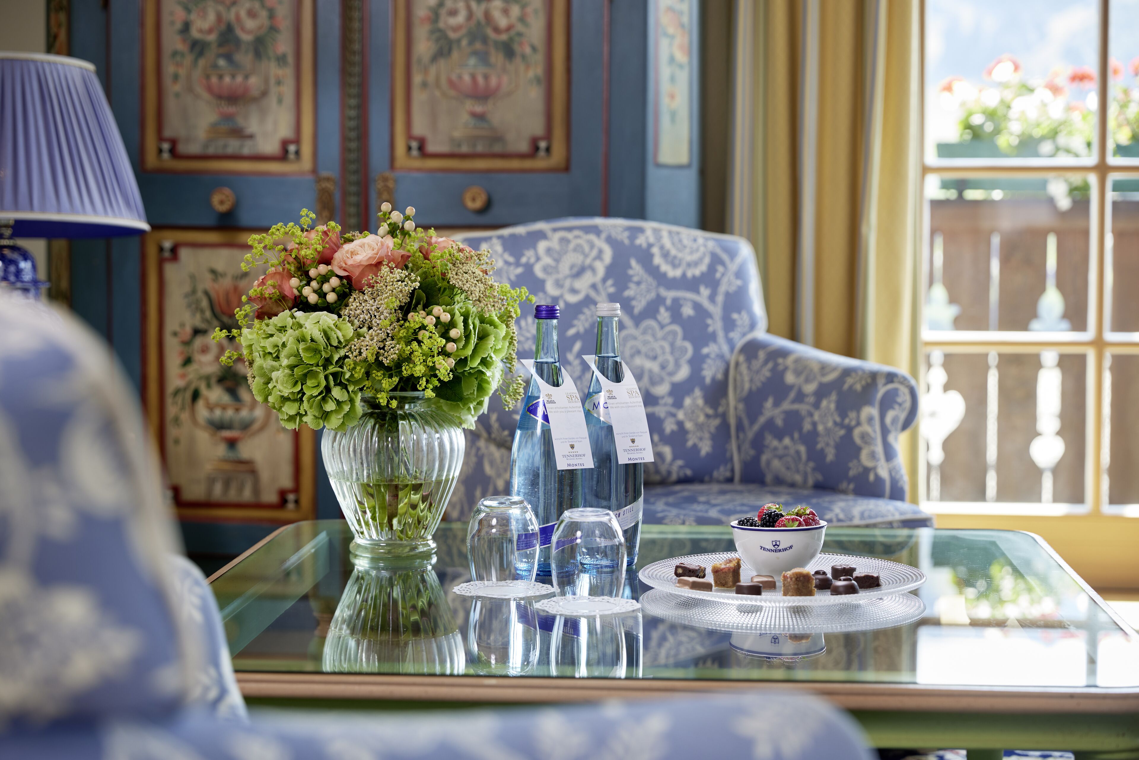 Una mesa con un arreglo floral, botellas y una bandeja de postres en una habitación con papel tapiz estampado y una ventana con vistas.