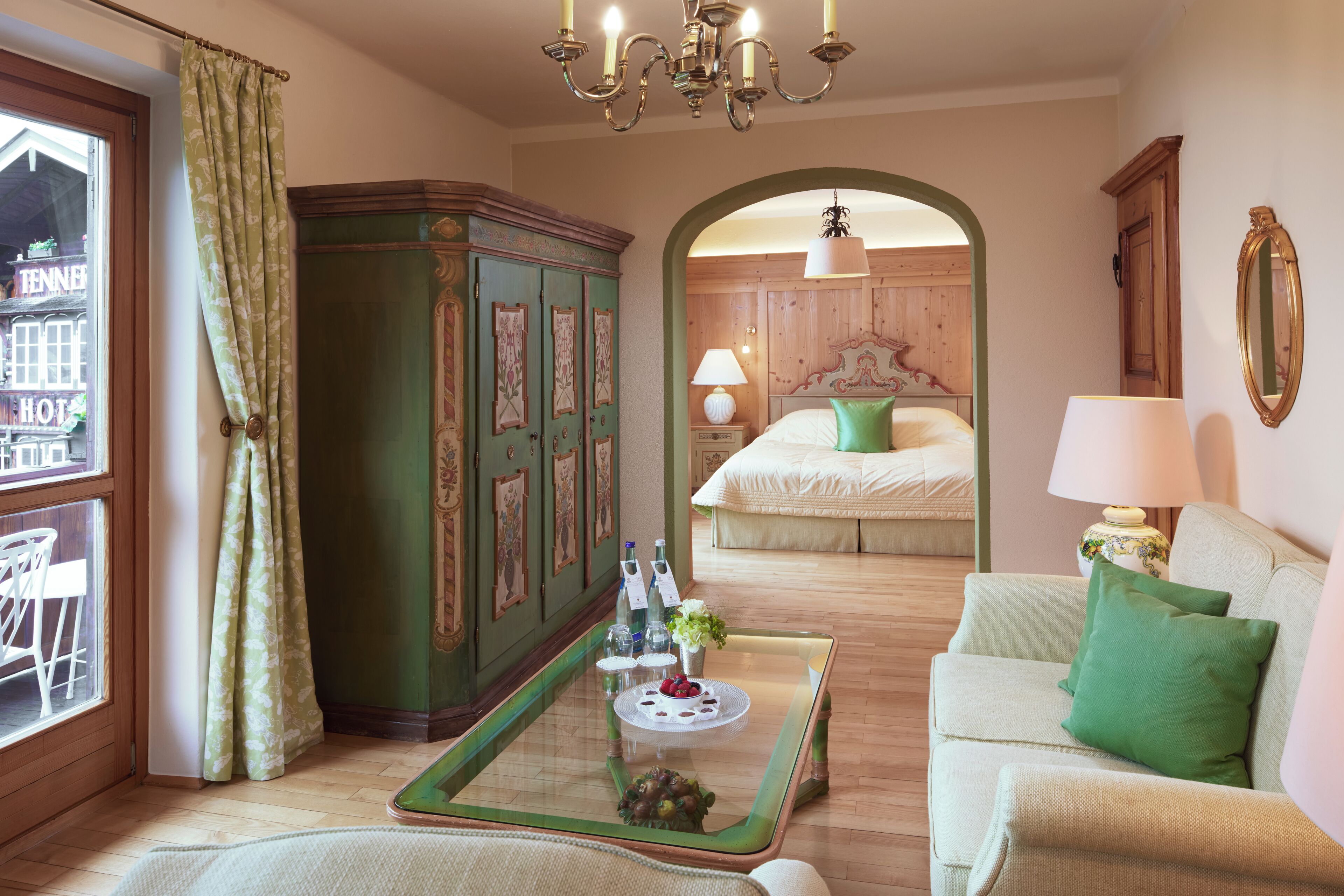 A hotel room with a bed, a sitting area with a sofa, a dining table with a tray, a green cabinet, and a view of a building outside.