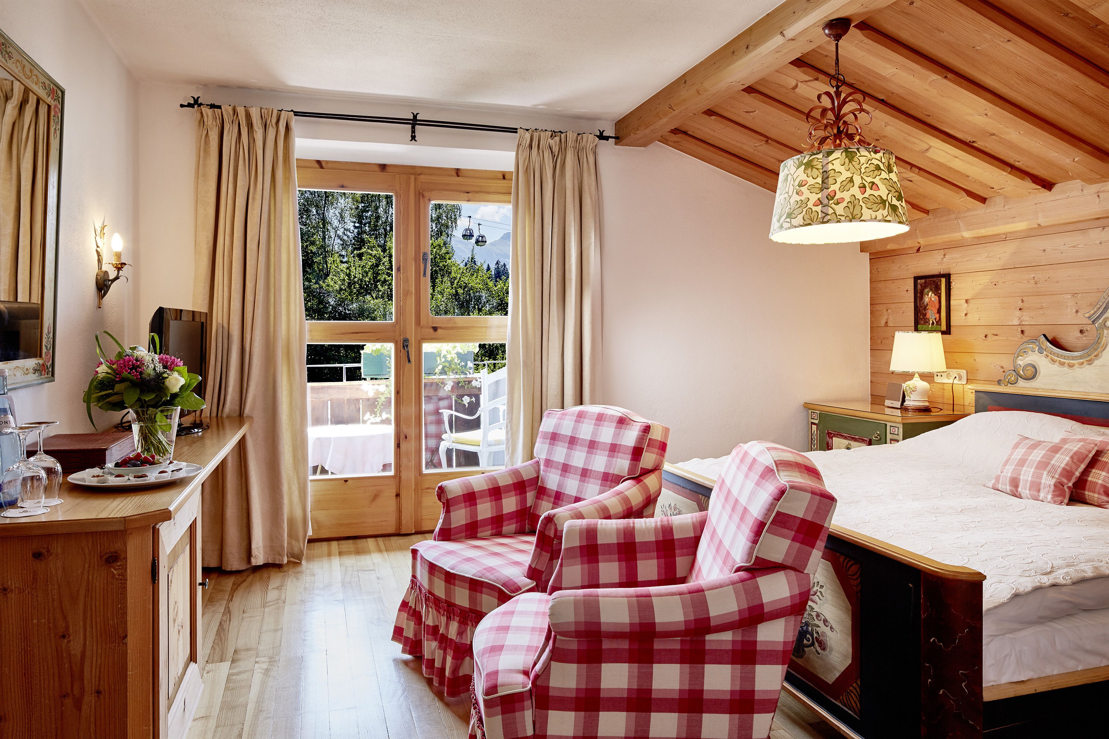 Une chambre confortable avec un plafond en bois, un lit recouvert d’une couette blanche, deux fauteuils à motifs à carreaux, une commode en bois et une fenêtre avec des rideaux.