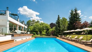 A swimming pool with wooden deck and lounge chairs, surrounded by trees and a modern building with large windows. - Relais & Châteaux Hotel Tennerhof (Kitzbuhel)