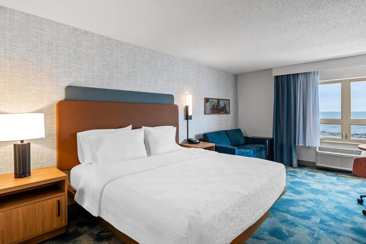 Una habitación de hotel con una cama grande, un escritorio con lámpara, una silla, una mesita, un cuadro en la pared y una ventana con cortinas.