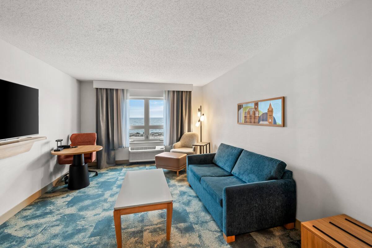 A hotel room with a large flat-screen TV, a sofa, a coffee table, a chair, a small table, a lamp, a painting, and a view of the sea.