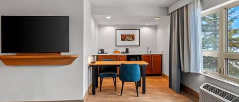 A modern living room with a flat-screen TV, a dining area with a wooden table and chairs, a kitchen with wooden cabinets, and a large window with curtains.