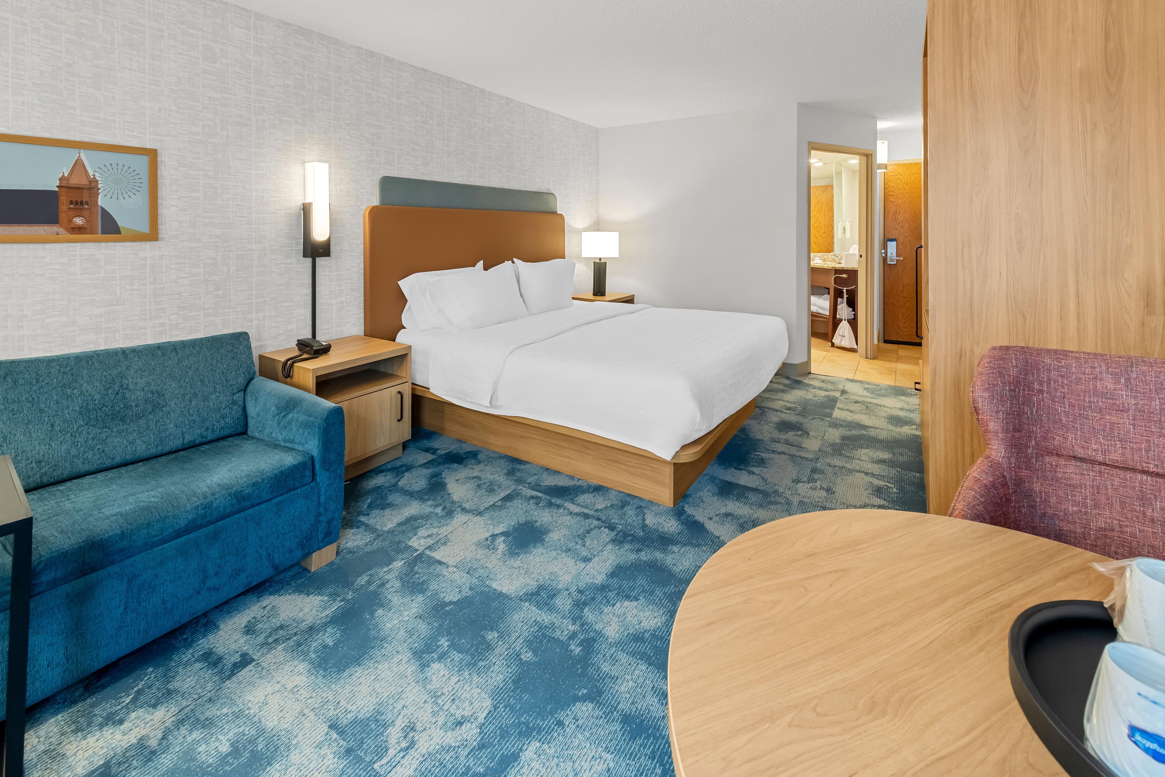 Una habitación de hotel con cama, sofá azul, mesa de madera y mesita de noche con lámpara.