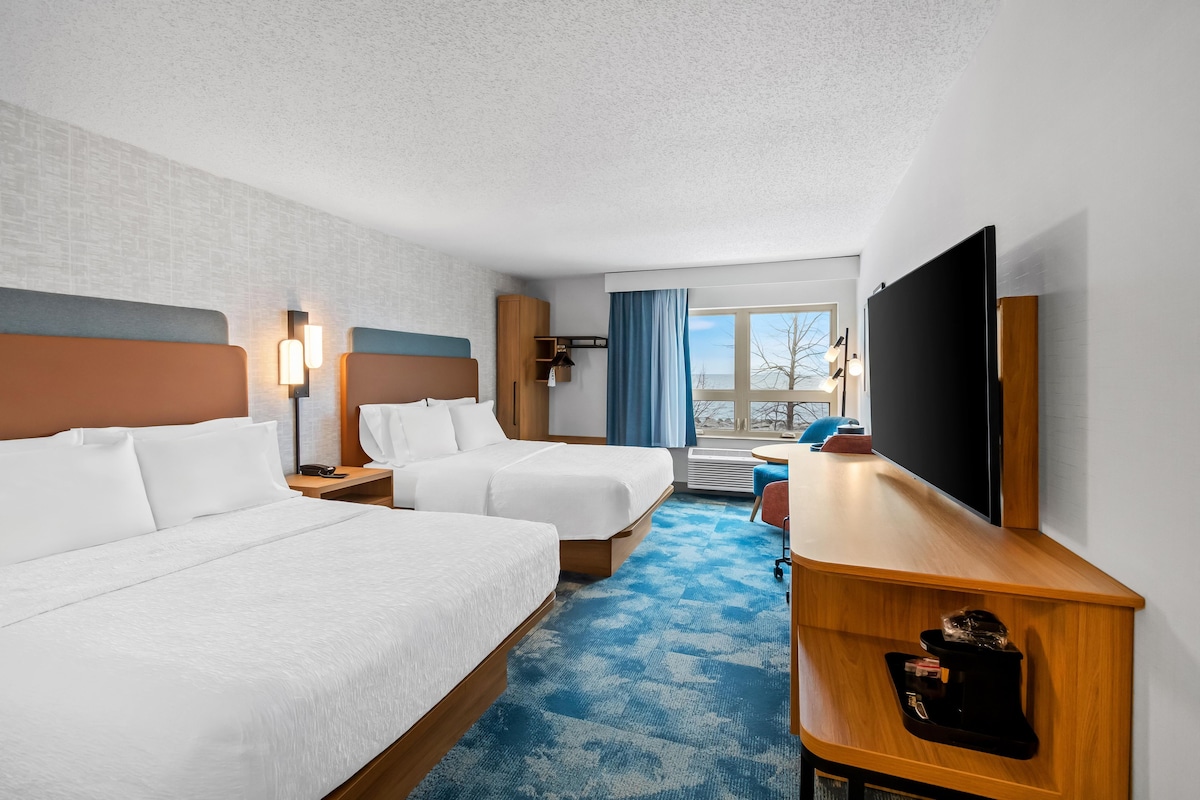 A hotel room with two beds, a desk, a TV, and a window with a view.
