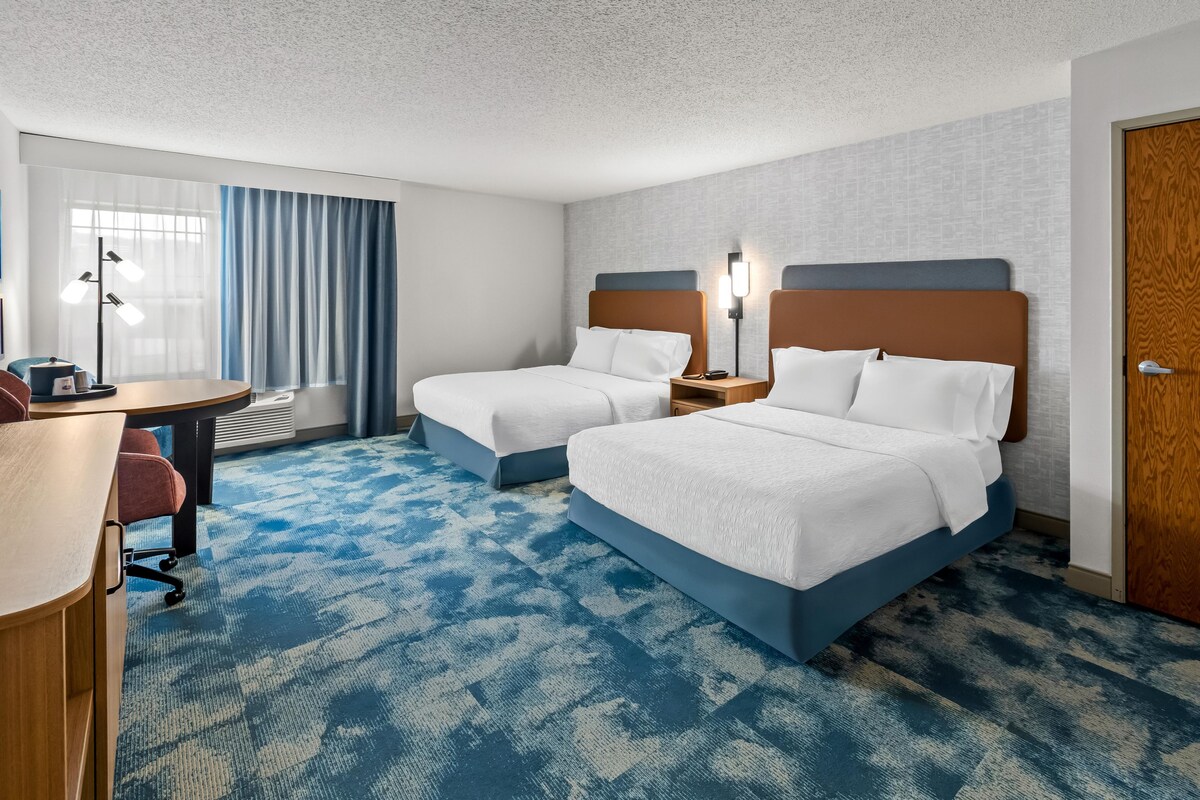 A hotel room with two beds, a desk, a chair, and a large window with curtains.