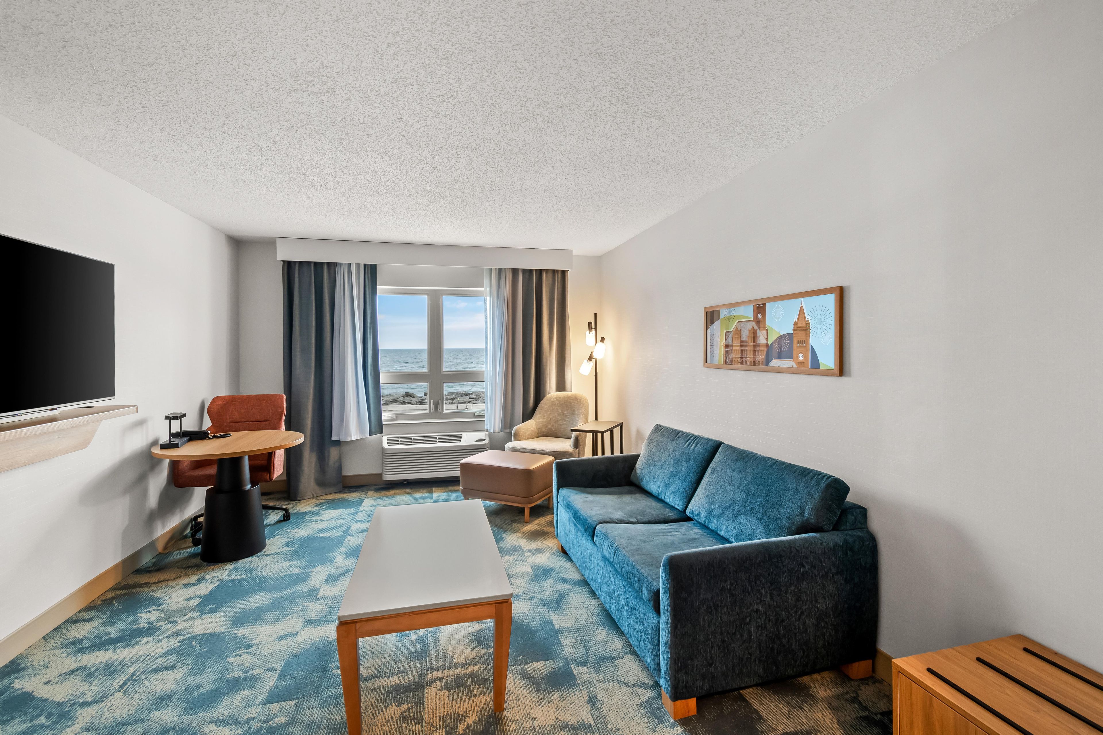 Habitación de hotel con sofá, una mesa, televisor, silla, lámpara y un cuadro.