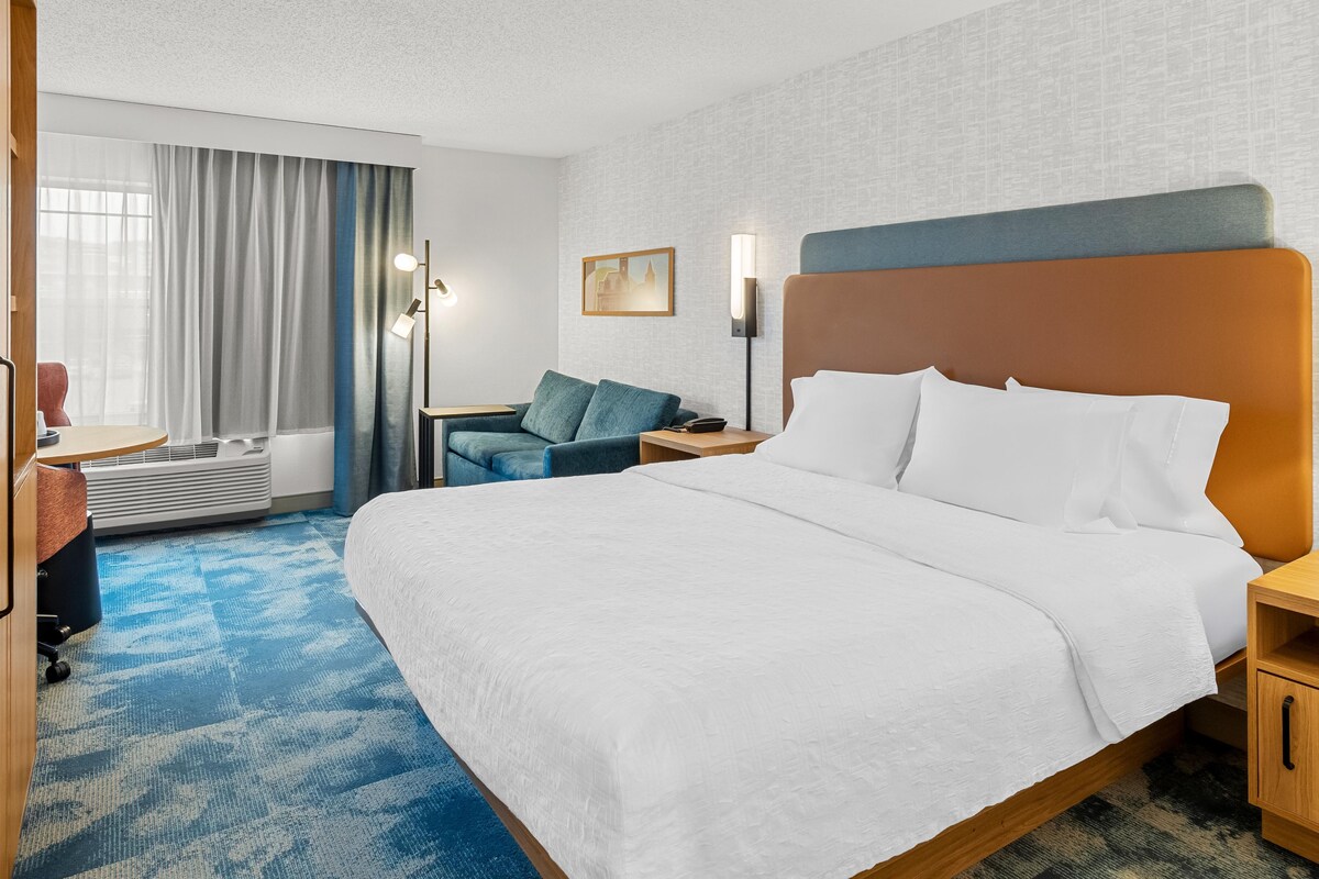 A hotel room with a large bed, a desk, a chair, a sofa, a nightstand, and a wall-mounted lamp.